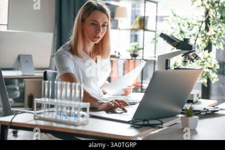 Utilisation d'un ordinateur portable. Jeune femme médecin en manteau est assise dans la clinique et travaille. Banque D'Images