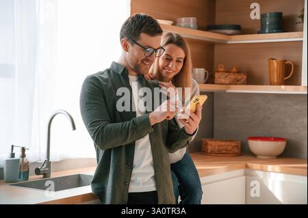 Homme montrant des médias à la femme au téléphone. Jeune couple est sur la cuisine ensemble. Banque D'Images