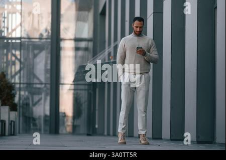 Bel homme marche, tenant un smartphone. À l'extérieur, près du bâtiment d'affaires. Banque D'Images