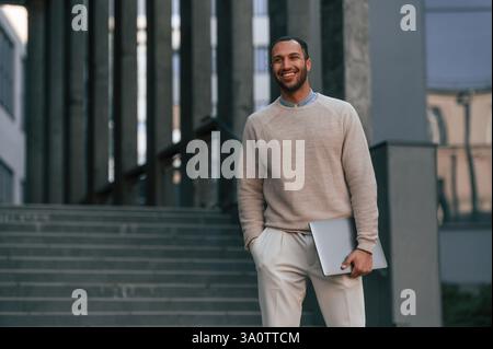 Positif bel homme noir est à l'extérieur près du bâtiment d'affaires tenant l'ordinateur portable. Banque D'Images