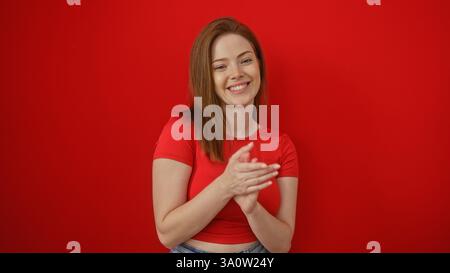 Jeune femme rousse applaudissant isolée sur un fond rouge avec un sourire joyeux, portant un haut rouge décontracté. Banque D'Images