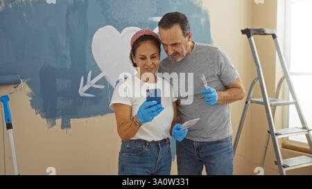 Couple hispanique peignant leur nouvelle maison, souriant dans le salon tout en tenant un téléphone, entouré d'outils de peinture, exprimant l'amour et créativit Banque D'Images