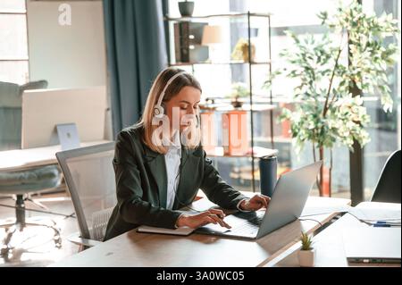 Dans les écouteurs, assis à table avec ordinateur portable. Jeune belle femme en vêtements formels travaille dans le bureau. Banque D'Images