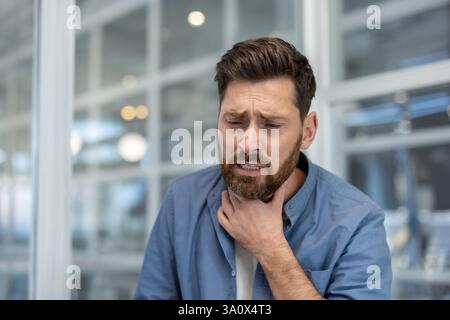 Un homme inquiet vêtu d'une chemise bleue décontractée tient son cou exprimant l'inconfort et la maladie, souffrant peut-être d'un mal de gorge. Banque D'Images