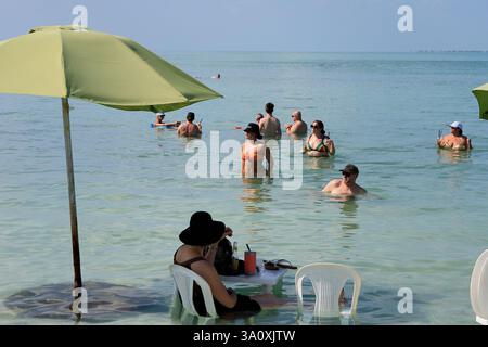 Un visiteur ayant un verre à table dans l'eau avec un groupe de touristes se rafraîchissent eux-mêmes dans l'eau de l'océan en arrière-plan. Secret Beach, près de San Pedro. Ambergris Caye, Belize Banque D'Images