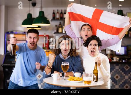Groupe d'amis, supporters sportifs acclamant l'équipe favorite avec drapeau de l'Angleterre tout en regardant le match à la télévision dans le bar Banque D'Images