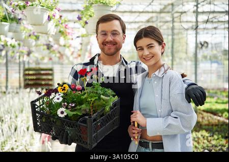 Sourire sincère, debout, tenant des fleurs. L'homme et la femme sont ensemble dans une serre lumineuse. Banque D'Images