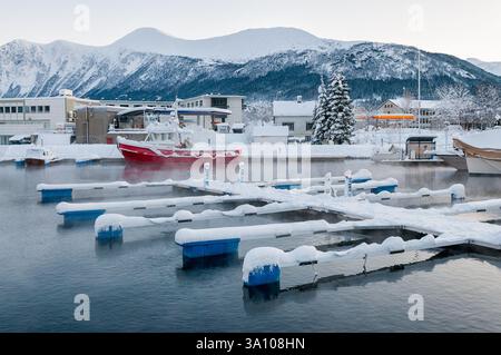La neige couvre la marina dans une ville calme du nord, avec des bateaux reposant doucement sur l'eau gelée et les montagnes en arrière-plan. Banque D'Images