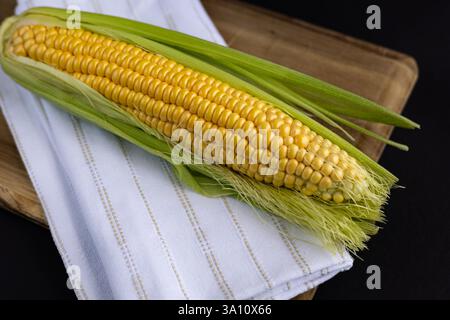 Épi de maïs frais avec des noyaux dorés vibrants partiellement enveloppé dans une balle verte placée sur une planche à découper en bois avec une serviette en lin blanc Banque D'Images
