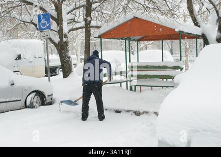 Une personne vêtue d'un vêtement d'hiver pelletant la neige d'un espace de stationnement désigné pour handicapés près d'un banc de parc enneigé. Un panneau bleu de stationnement pour handicapés Banque D'Images