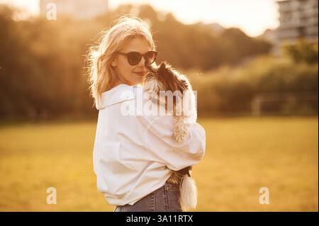 Femme en lunettes de soleil est debout sur le champ d'été et tenant petit chien. Banque D'Images