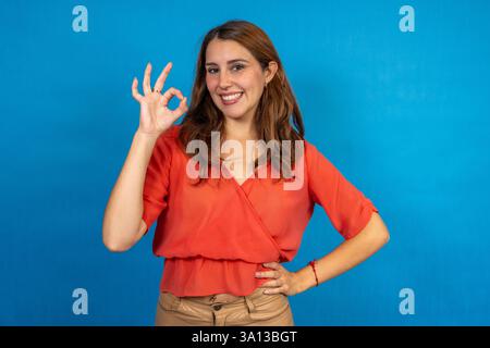 Femme d'affaires souriante faisant un geste OK avec sa main sur un fond bleu Banque D'Images