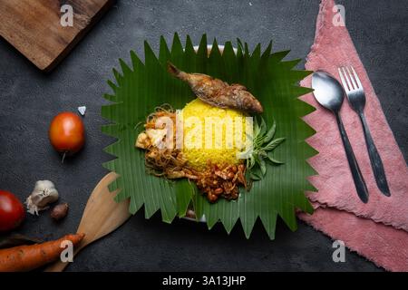 Authentique Nasi Kuning indonésien servi sur des feuilles de bananier avec du poisson frit, des vermicelles, du tempeh, des arachides et des herbes fraîches. Banque D'Images