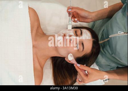 Application d'un masque facial en argile. Visage de femme obtenant des soins du visage par des mains esthéticiennes au salon de spa. Banque D'Images