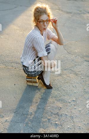Jeune femme nerdy dans des lunettes surdimensionnées peine à transporter des livres lourds, bâillant ou haletant dans l'épuisement tout en se tenant dehors à la lumière du soleil. Banque D'Images
