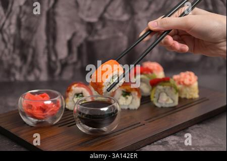 La main de la femme tenant le rouleau de sushi a utilisé des baguettes et trempé dans la sauce soja. Ensemble de rouleau de sushi sur une planche de service en bois. Fond sombre Banque D'Images