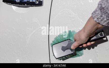 Une main lavant le côté d'une voiture blanche à l'aide d'une brosse et d'eau savonneuse Banque D'Images