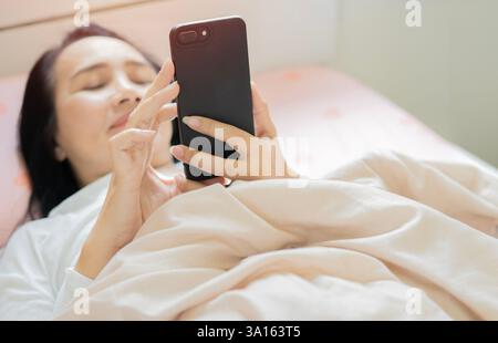 Sommeil sain, heureuse femme d'âge moyen détendue avec téléphone portable couché sur le lit sur un oreiller confortable doux blanc dans la chambre à coucher à la maison Banque D'Images