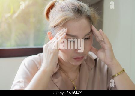 Adulte belle femme asiatique assise près de la fenêtre et souffrant de maux de tête désespérée, stressée à cause de la douleur et de la migraine. Banque D'Images