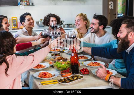Des amis multiethniques joyeux grillent avec du vin rouge tout en déjeunant ensemble à la maison Banque D'Images