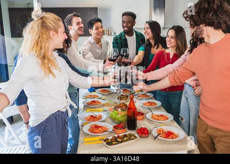 Groupe d'amis joyeux multi-ethniques grillant des verres de vin rouge tout en dégustant un déjeuner spaghetti à la maison Banque D'Images