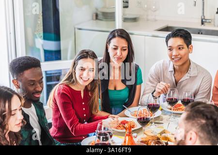 Groupe de jeunes amis multiethniques dégustant un repas de pâtes avec sauce tomate et vin rouge à la maison Banque D'Images