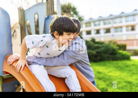 Grand-père tenant petit-fils glissant sur un toboggan orange à l'aire de jeux, s'amusant ensemble par une journée ensoleillée Banque D'Images