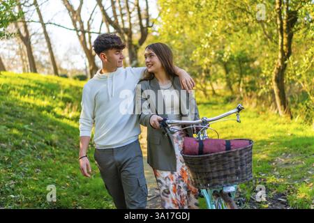 Jeune couple souriant marchant à vélo dans un parc ensoleillé, entouré d'arbres verdoyants, partageant un moment joyeux ensemble Banque D'Images