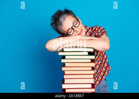 Femme âgée souriante appuyée sur une pile de livres, portant des lunettes et une chemise vibrante sur un fond bleu Banque D'Images