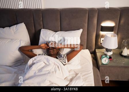 Femme noire se reposant dans le lit, les mains protégeant les yeux, niché parmi des oreillers moelleux, table de chevet tenant réveil et lampe, représentant la détente paisible matin ou nocturne Banque D'Images