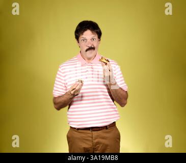 Jeune adulte aux cheveux foncés avec moustache, polo rayé rose et blanc, pantalon brun, regardant la caméra et mangeant un gâteau avec une fa satisfaite Banque D'Images