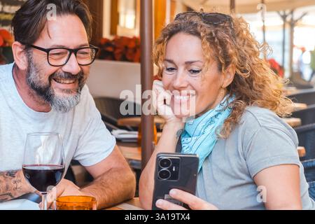 Couple caucasien lisant la notification téléphonique avant le déjeuner assis à la table du restaurant ensemble s'amusant. Les gens homme et femme regardant les commentaires d'examen ou le menu en ligne choisir quoi manger Banque D'Images