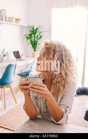 Femme blonde bouclée assise à la table de la cuisine buvant un café regarde et pense tout en se relaxant. La femme d'âge moyen se détend en prenant une pause du travail. Banque D'Images