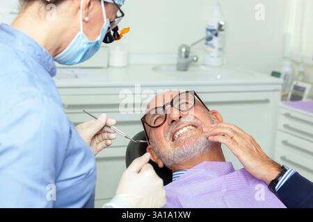 Dentiste portant une loupe et un masque chirurgical examinant un patient âgé souffrant de maux de dents dans une clinique dentaire brillante Banque D'Images