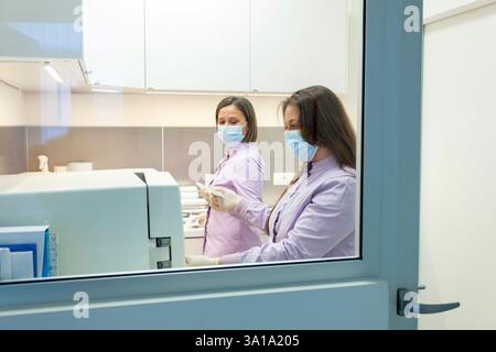 Deux dentistes travaillent avec un stérilisateur autoclave dans une clinique dentaire, portant des masques chirurgicaux et des gants, assurant l'hygiène et la sécurité Banque D'Images