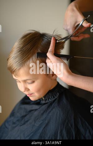 Le coiffeur amincit les cheveux du garçon avec des ciseaux amincissants. Banque D'Images