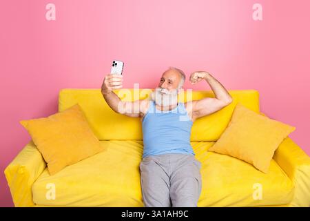 Homme mûr avec la barbe fléchissant bras et souriant tout en prenant selfie sur smartphone d'un canapé jaune sur fond rose à l'intérieur Banque D'Images