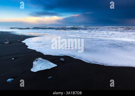 Vue sur les vagues de la mer qui coulent autour des blocs de glace au coucher du soleil sur la célèbre plage Diamond. Jökulsarlon, Diamond Beach, Austurland, Islande, Europe du Nord. Banque D'Images