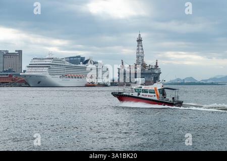 Un petit bateau naviguant sur les eaux bleues pittoresques de la baie de Guanabara avec MSC Orchestra et une plate-forme pétrolière amarrée à l'embarcadère de Mauá sous un ciel ensoleillé et nuageux d'été. Banque D'Images