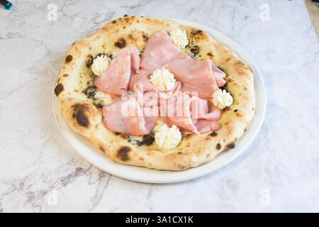 Une pizza avec jambon et fromage sur une assiette blanche. Le jambon est coupé en petits morceaux et le fromage est réparti uniformément sur la pizza. La pizza est placée dessus Banque D'Images