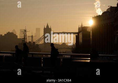 Vue tôt le matin des navetteurs marchant sur London Bridge avec vue sur Tower Bridge et HMS Belfast silhouetté par le soleil tôt le matin. Banque D'Images