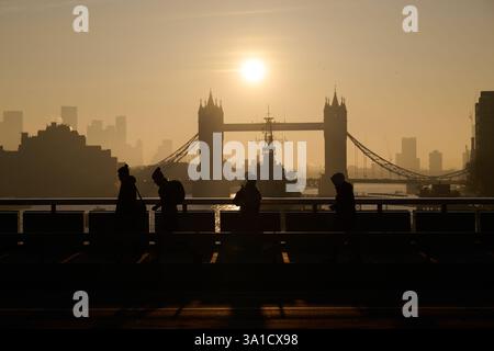Vue tôt le matin des navetteurs marchant sur London Bridge avec vue sur Tower Bridge et HMS Belfast silhouetté par le soleil tôt le matin. Banque D'Images