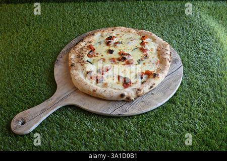 Une délicieuse pizza avec du fromage et des garnitures est assis sur une planche de bois sur une pelouse verte luxuriante. La pizza est placée sur une planche de bois qui est sur un vert Banque D'Images