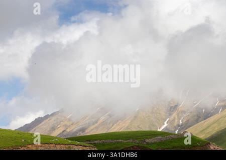 Un paysage de montagne pittoresque partiellement couvert de nuages brumeux. Les collines verdoyantes contrastent avec les pentes rocheuses et les parcelles de neige. Un ciel vaste avec doux, blanc Banque D'Images