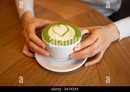 Mains de femme tenant une tasse blanche de latte matcha décorée d'un art latte en forme de cœur, placée sur une table en bois, créant une atmosphère chaleureuse et accueillante Banque D'Images