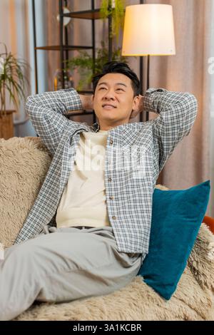 Heureux homme chinois d'âge moyen insouciant assis confortablement avec la main derrière la tête sur le canapé dans le salon à la maison. Souriant mec coréen se relaxant sur le canapé pensant regarder autour dans la chambre de l'appartement. Banque D'Images
