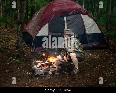 Homme agenouillé à côté d'un feu de camp dans la forêt tout en campant avec une tente pendant une journée pluvieuse du matin portant short et imperméable Banque D'Images