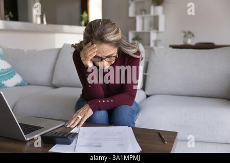 Une femme âgée stressée est assise à table avec des factures et un ordinateur portable Banque D'Images