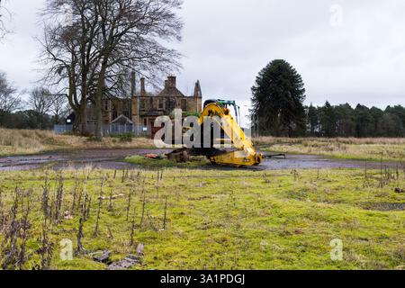 Excavatrice jaune abandonnée dans un champ herbeux devant un asile psychiatrique de l'hôpital de Hartwood - Maison du surintendant médical abandonnée Banque D'Images