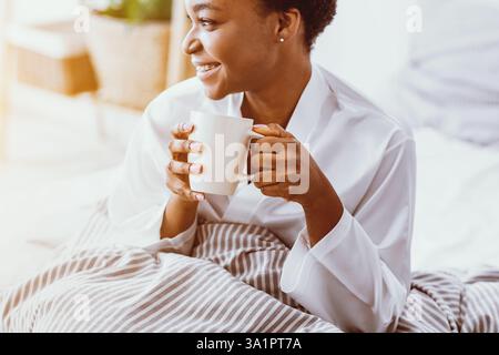 Café du matin. Heureuse fille afro-américaine regarde loin et tient la tasse Banque D'Images